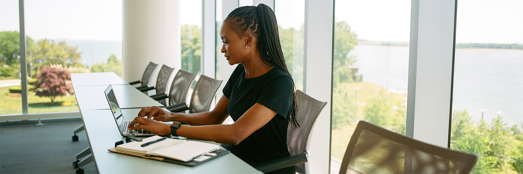 A UMass Boston student wearing a black dress sits at a table with her laptop in front of windows with a water view