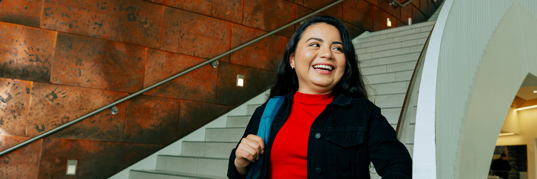 Student smiling holding backpack in front of university hall inner staircase in lobby.