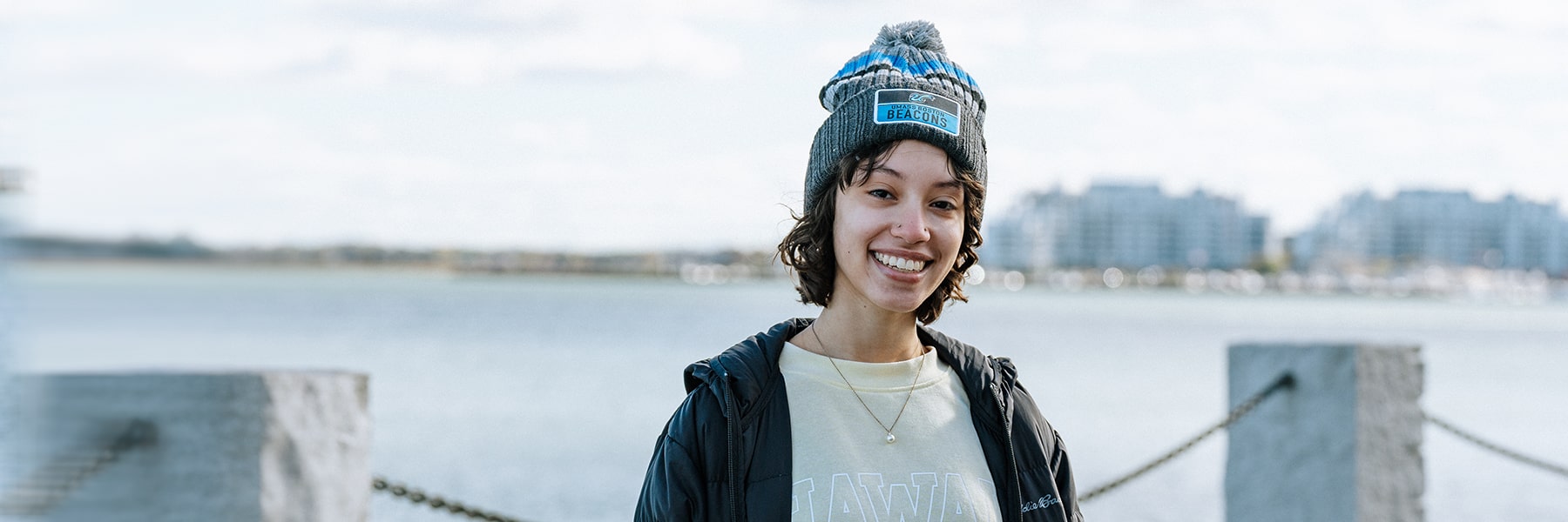 Student wearing a beanie posting by the water on the Harborwalk.