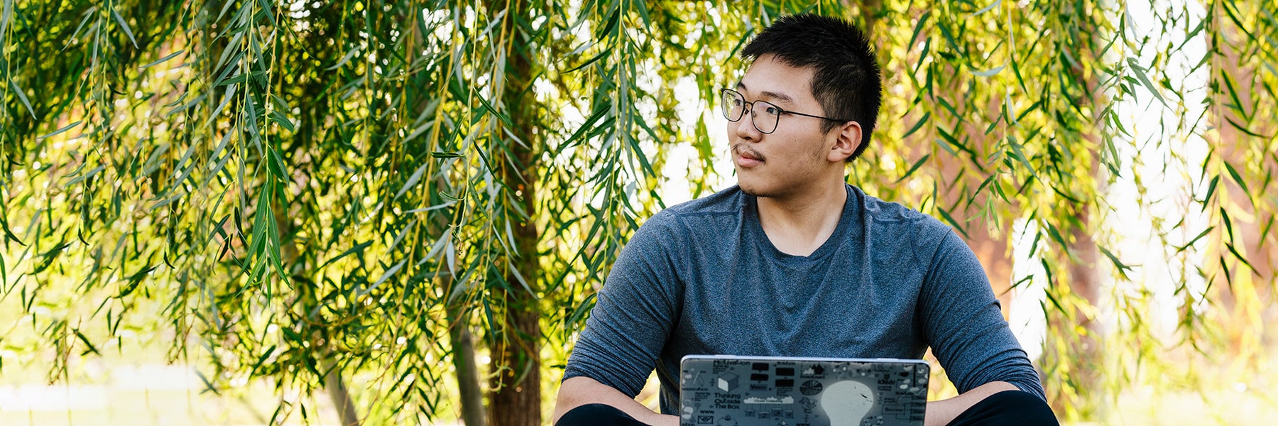 Student works on laptop under trees.